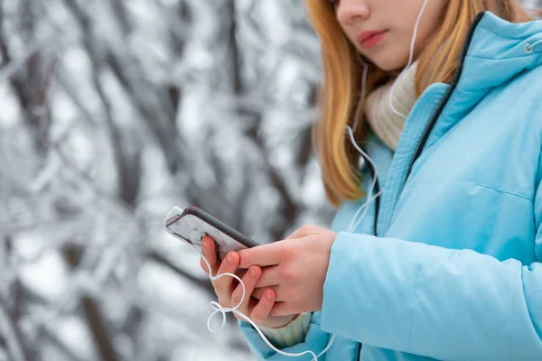 Nahaufnahme von erfrorenen Händen eines jungen hübschen Mädchens, das in einem Winter-Snowpark spazieren geht und sich online Videos mit einem Smartphone ansieht oder Musik mit Kopfhörern hört — Stockfoto