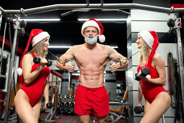 Різдво. М'язовий красень Санта Клаус з сірою бородою і голим торсом тренується в спортзалі разом з двома сексуальними дівчатами в спортивних купальниках і капелюшках з кришталевими капелюхами, що тримають парасольки. — стокове фото