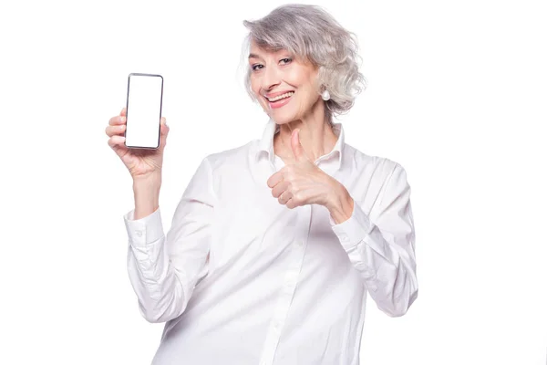 Eine schöne, fröhliche, moderne Seniorin blickt mit einem breiten Lächeln in die Kamera, zeigt ein Smartphone in der erhobenen Hand und zeigt den Daumen in die Höhe, um ihre Zustimmung auszudrücken, isoliert auf weißem Hintergrund. — Stockfoto