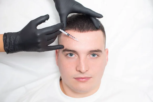 Zbliżenie twarzy młodego mężczyzny i rąk kosmetologa w czarnych rękawiczkach medycznych wstrzykujących botoks z zmarszczek do miejsc oznaczonych białym ołówkiem na czole pacjenta — Zdjęcie stockowe