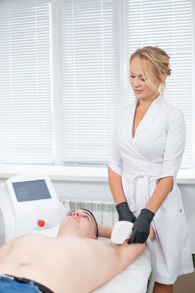 A pretty cosmetologist at work in her modern cosmetology office during the procedure of laser hair removal from the armpit of a young man — Stock Photo, Image