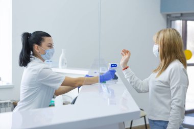 Modern bir hastanenin resepsiyon masası çalışanı tarafından kadın vücut ısısının temassız termometreyle ölçüldüğü bir fotoğraf. Yayılmamaları için alınan önlemlere uyma