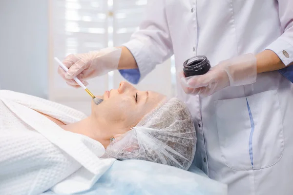 Widok boczny dłoni specjalisty kosmetologii stosującego nanogel węglowy na twarz młodej pacjentki za pomocą pędzla, przygotowującego się do zabiegów obierania węgla — Zdjęcie stockowe