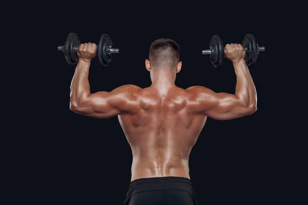 Vista posterior cercana del cuerpo muscular y manos fuertes levantando pesadas mancuernas aisladas sobre fondo negro — Foto de Stock