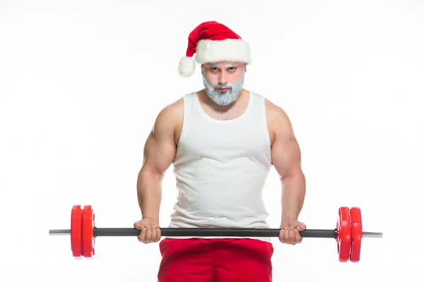 Navidad. Santa Claus musculoso fuerte con la barba gris que usa el sombrero de Navidad y los pantalones cortos rojos está levantando una barra pesada en un gimnasio que realiza un entrenamiento aislado en fondo blanco. — Foto de Stock