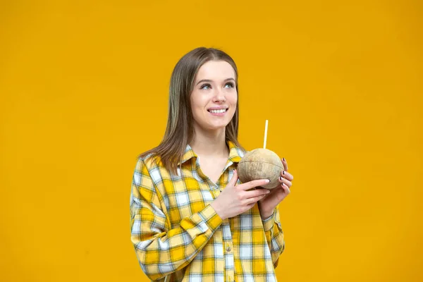 Tourné d'une jolie jeune femme souriante regardant de côté tenant dans les deux mains de la noix de coco avec une paille isolée sur fond jaune. Concept de mode de vie sain, bonne nutrition, régime alimentaire, végétalien — Photo