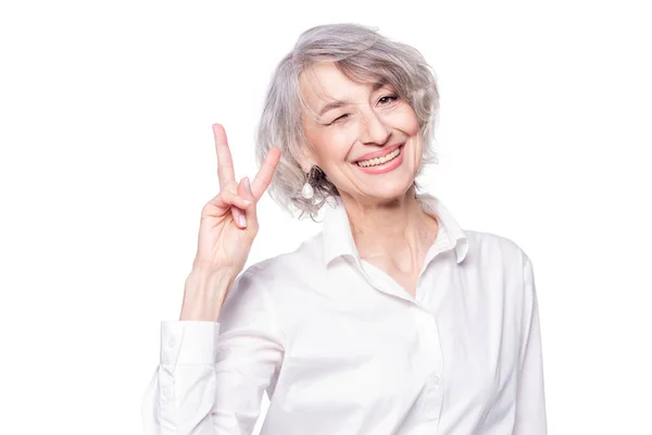 Nahaufnahme Porträt einer älteren grauhaarigen Frau mit elegantem Hemd, die vor isoliertem weißem Hintergrund steht, lächelnd mit glücklichem Gesicht in die Kamera zwinkert und das Siegeszeichen macht — Stockfoto
