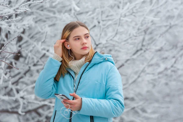 Μπροστά όψη ενός όμορφου έφηβου κοριτσιού ακούγοντας μουσική με ακουστικά σε ένα smartphone ενώ περπατούσε σε ένα χιονισμένο χειμερινό πάρκο κοιτάζοντας στην άκρη και φτιάχνοντας τα μαλλιά της — Φωτογραφία Αρχείου