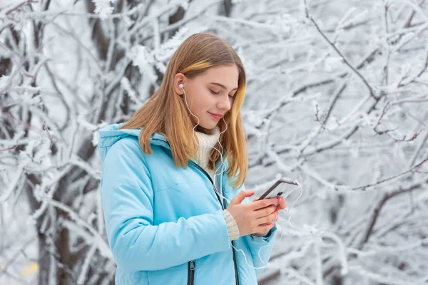 Närbild skott av frysta frostbitna händer av en ung söt flicka som går i en vinter snö park titta på videor på nätet i en smartphone eller lyssna på musik med hörlurar — Stockfoto