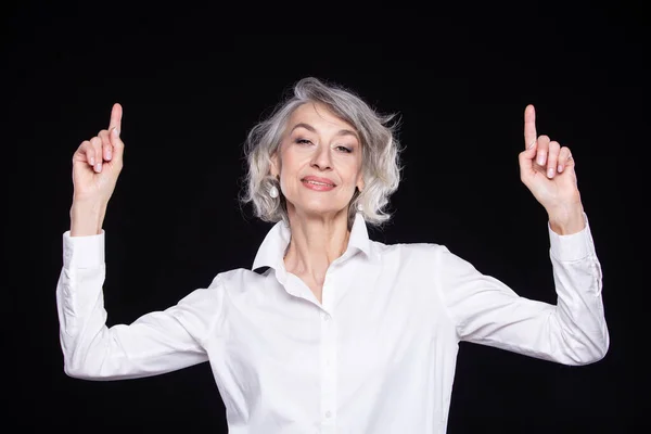 Portrait of a modern happy senior woman pointing upwards with two hands, isolated on a black background — Stock Photo, Image