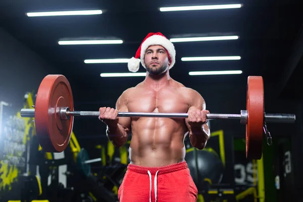 Передній вид сексуального м'язистого чоловіка в різдвяному капелюсі і червоні шорти, що піднімають штангу в спортзалі — стокове фото