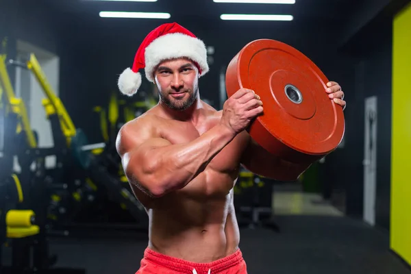 Молодий сильний м'язовий Санта Клаус у червоному капелюсі та короткометражках дивиться на камеру, яка піднімає важкий диск у спортзалі. — стокове фото