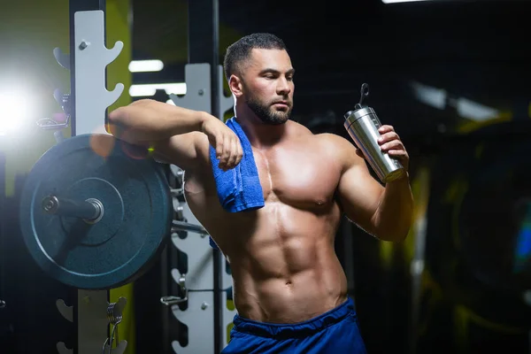 Young sexy sportsman is relaxing after a hard training in a gym standing near a barbell with a towel and drinking water — Stock Photo, Image