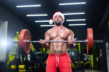Noel şapkalı ve kırmızı şortlu seksi kaslı bir adamın spor salonunda halter kaldırışının ön görüntüsü.