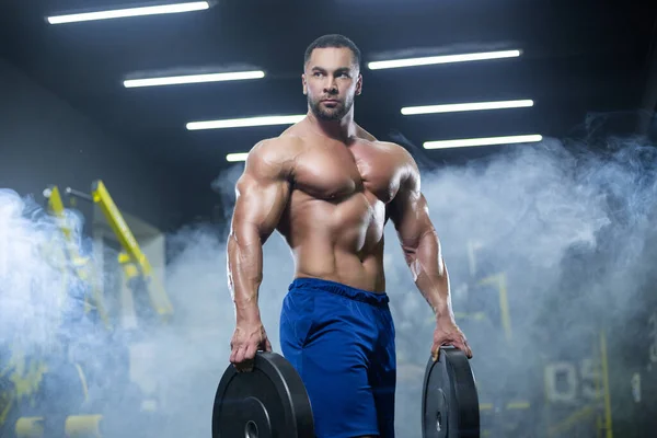 Hombre atleta musculoso guapo está de pie en un gimnasio contra el fondo de humo sosteniendo discos de barra en ambas manos —  Fotos de Stock