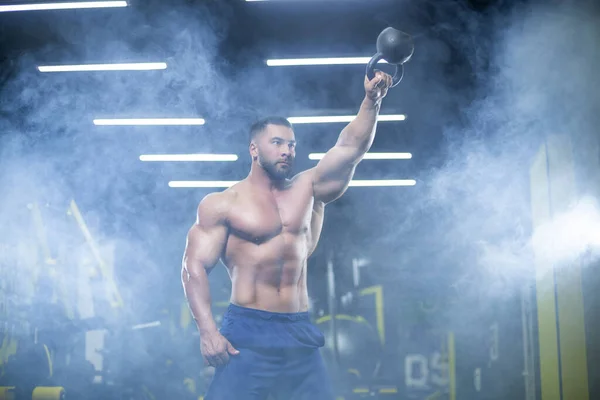 Portrait of a muscular sportsman performing kettlebell swings with one hand in a gym standing in smoke — Stock Photo, Image