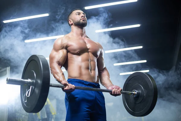 Vista de abajo hacia arriba de un atleta muscular levantando pesadas barras mostrando sus músculos en un gimnasio parado en el humo — Foto de Stock