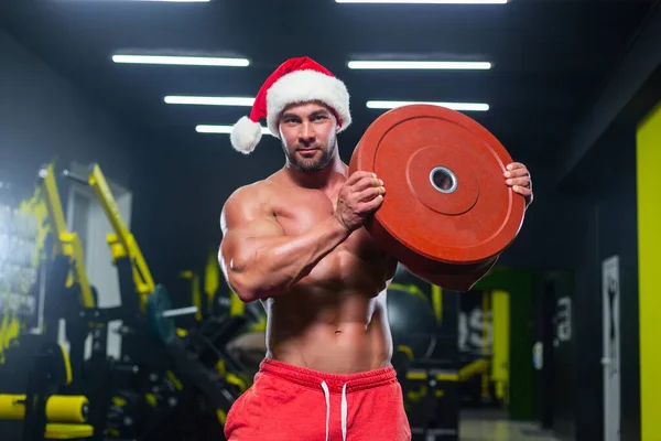 Молодий сильний м'язовий Санта Клаус у червоному капелюсі та короткометражках дивиться на камеру, яка піднімає важкий диск у спортзалі. — стокове фото