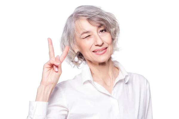 Ritratto ravvicinato della donna anziana dai capelli grigi che indossa una camicia elegante in piedi su uno sfondo bianco isolato sorridente con volto felice che strizza l'occhio alla macchina fotografica facendo segno di vittoria — Foto Stock