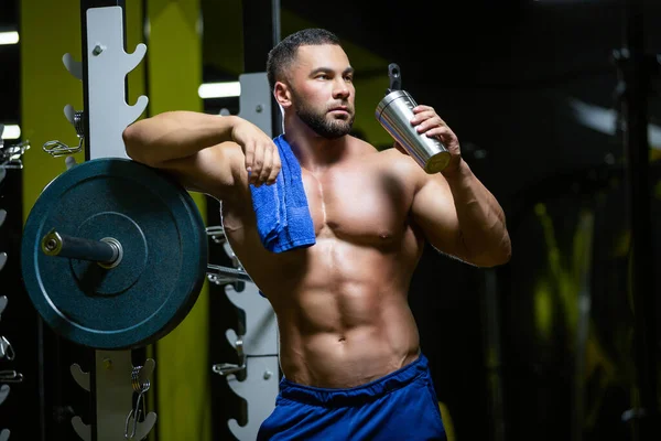 Jonge sexy sportman is ontspannen na een harde training in een fitnessruimte in de buurt van een lange halter met een handdoek en drinkwater — Stockfoto