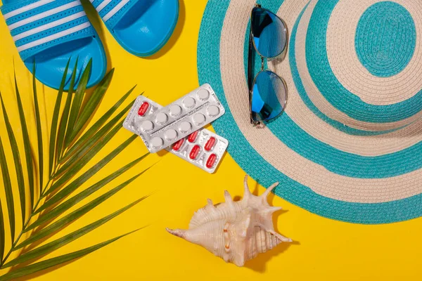Widok z góry na białe i czerwone tabletki w blistrze, niebieski kapelusz, niebieskie japonki, okulary przeciwsłoneczne, muszla morska, liść palmowy na żółtym tle. Koncepcja wypoczynku na plaży, wycieczka po morzu, ciepłe słoneczne lato. Aklimatyzacja — Zdjęcie stockowe