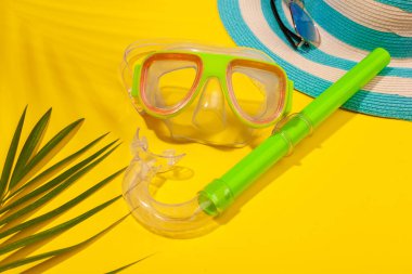 Sarı arkaplanda plaj aksesuarları, güneş gözlükleri, çizgili mavi şapka, dalgıç maskesi ve palmiye yaprağı gölgesinde palmiye yaprağı. Uzun zamandır beklenen deniz ve seyahat tatili kavramı. Reklamcılık