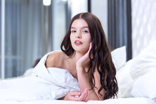 Eine attraktive junge Frau liegt nackt auf dem Bett unter der Decke und blickt lächelnd in die Kamera, den Kopf auf die Hand gestützt. — Stockfoto