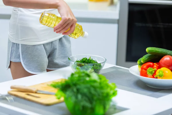 Foto de close-up de uma menina em shorts derramando óleo vegetal em uma salada de vitamina vegetal em uma tigela de vidro na cozinha — Fotografia de Stock