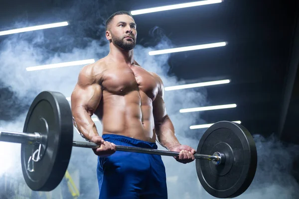 Vista de abajo hacia arriba de un atleta muscular levantando pesadas barras mostrando sus músculos en un gimnasio parado en el humo — Foto de Stock