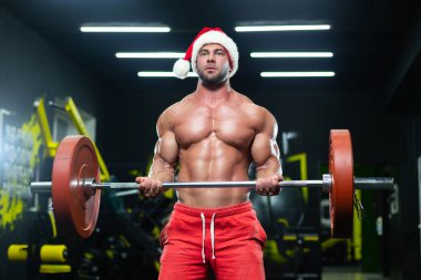 Noel şapkalı ve kırmızı şortlu seksi kaslı bir adamın spor salonunda halter kaldırışının ön görüntüsü.