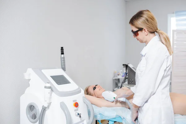 Foto de un dispositivo cosmetológico moderno y una dermatóloga femenina en gafas protectoras realizando un procedimiento de depilación láser de las axilas de una niña — Foto de Stock