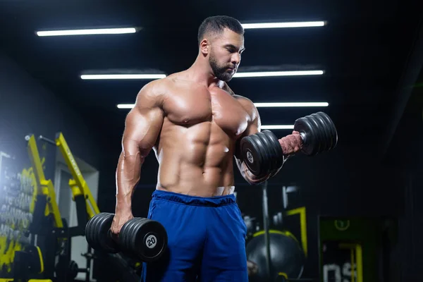 Front view of a muscular bodybuilder lifting dumbbells in blue shorts posing in a gym — 图库照片