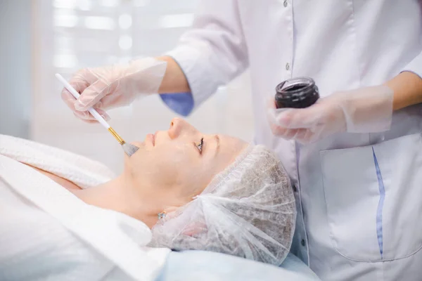 Widok boczny dłoni specjalisty kosmetologii stosującego nanogel węglowy na twarz młodej pacjentki za pomocą pędzla, przygotowującego się do zabiegów obierania węgla — Zdjęcie stockowe