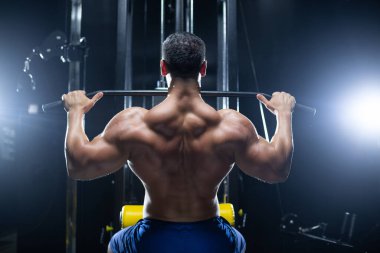 Yakışıklı fitness adamı spor salonunda, arka planda üst blok makinesini kullanarak egzersiz yapıyor.