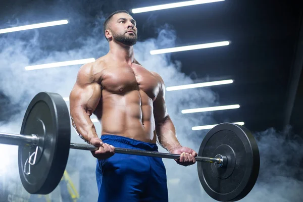 Bottom up view of a muscular athlete lifting heavy barbell showing his muscles in a gym standing in smoke — Stock Photo, Image