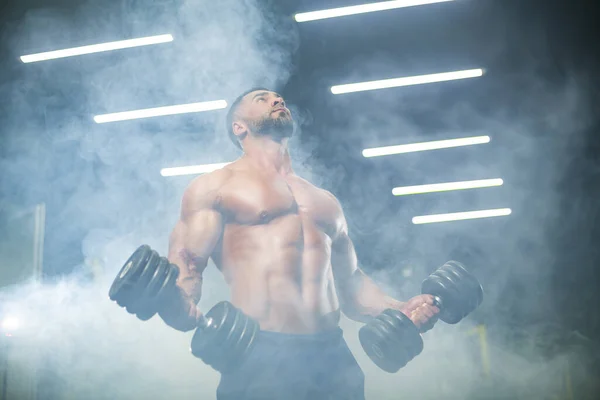 Bottom up view of a strong man athlete lifting dumbbells in the gym posing in smoke — Stock Photo, Image