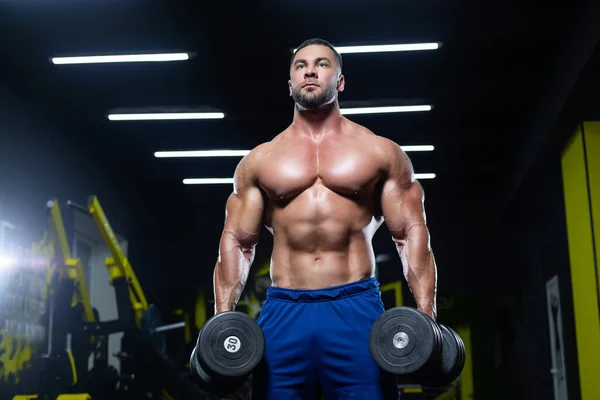 Front view of a muscular bodybuilder posing with dumbbells in blue shorts in a gym — Stock Photo, Image