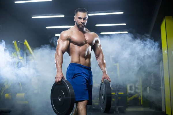 Hombre atleta musculoso guapo está de pie en un gimnasio contra el fondo de humo sosteniendo discos de barra en ambas manos — Foto de Stock