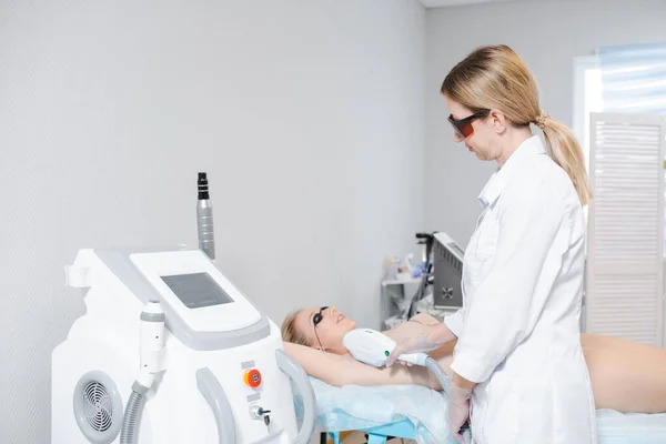 Foto de um moderno dispositivo de cosmetologia e uma dermatologista feminina em óculos de proteção realizando um procedimento de depilação a laser das axilas de uma jovem — Fotografia de Stock