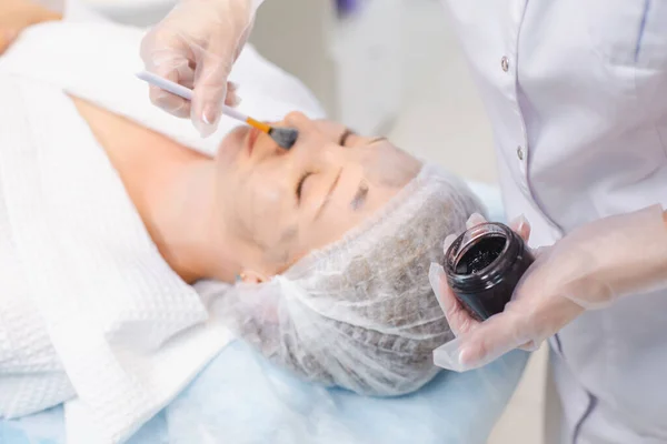 Hands of cosmetology specialist applying carbon nanogel on young woman patient face using brush, getting ready for carbon peeling procedures — Stock Photo, Image