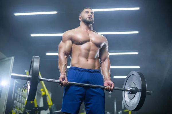 Снизу вверх вид мускулистого спортсмена, поднимающего тяжелую штангу, показывающего свои мышцы в спортзале — стоковое фото