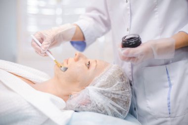 Genç hasta yüzüne karbon nanojel uygulayan kozmetoloji uzmanının elleri, fırça kullanarak karbon soyma prosedürlerine hazırlanıyor.