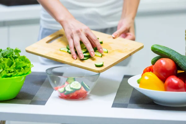 Primer plano de una chica manos rebanando verduras con un cuchillo en una tabla de cortar para una ensalada vegana de vitaminas vegetales y ponerlas en un tazón de vidrio mientras se cocina el desayuno en la cocina — Foto de Stock