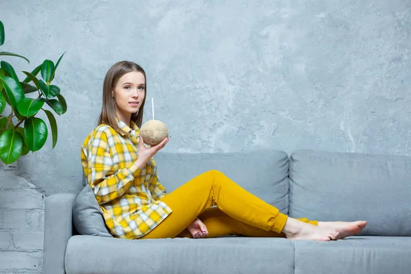 Foto de una hermosa joven sentada con las piernas en un sofá y sosteniendo en una mano un coco con una paja. Concepto de relajación, meditación, vegano, nutrición — Foto de Stock