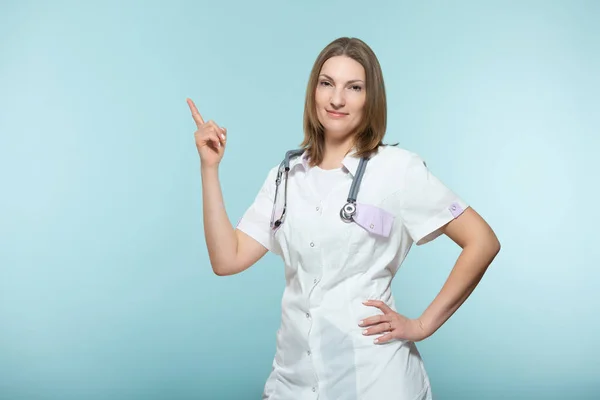 A beautiful female doctor with a stethoscope points to the place where there will be important information, on a blue background. Copy paste. healthcare concept. — Stock Photo, Image