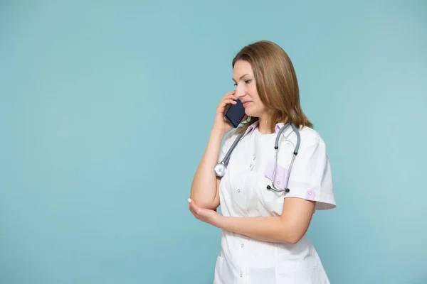 A woman doctor with a smartphone conducts an online consultation, supports the patient by phone, on a blue background. Copy paste. Distance healthcare concept. — Stock Photo, Image