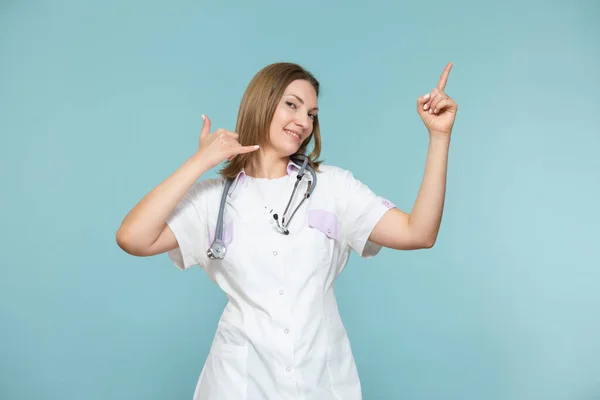 Una donna medico mostra un simbolo di un telefono e con la mano indica un metodo per la pubblicità o un numero di telefono, su uno sfondo blu. Copia incolla. concetto di assistenza sanitaria. — Foto Stock