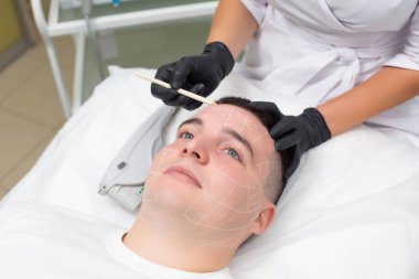 Siyah eldiven giymiş bir kozmetik uzmanının elleri beyaz bir kozmetik kalem tutarken, dermatoloji ofisinde kanepede yatan yakışıklı bir adamın portresi.