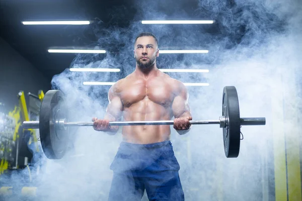 Close-up zicht op een sterke gespierde atleet tillen zware lange halter tonen zijn spieren in een sportschool staande in rook — Stockfoto