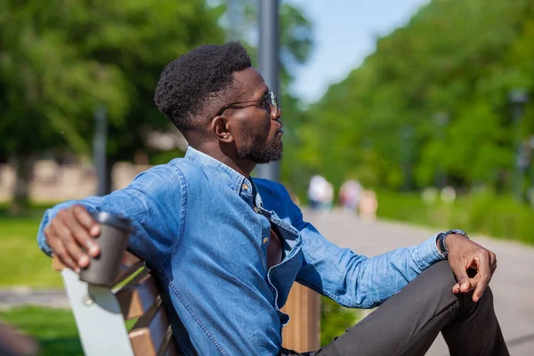 Kendine güvenen Afro-Amerikan bir iş adamının portresi şık giysiler içinde şehir parkında bankta oturuyor sıcak kahve içiyor ve kameraya bakıyor. — Stok fotoğraf